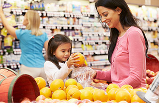 Small child putting orange in bag held by mother in produce aisle at grocery store